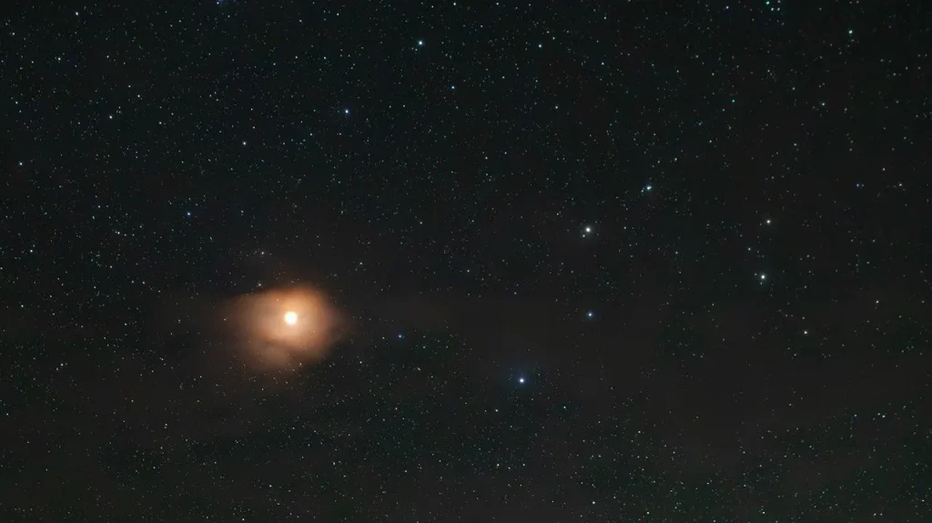Astrophotograph showing the bright orange-red star Antares surrounded by a glowing amber nebula against a star-filled night sky, with a vivid blue star visible nearby to the lower right.