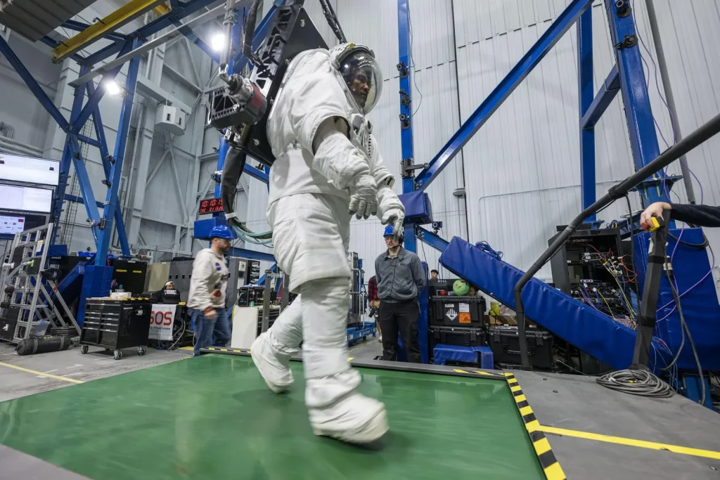 Artemis II astronaut Victor Glover walks on a treadmill while in a space suit harnessed to NASA’s Active Response Gravity Offload System at NASA’s Johnson Space Center. Glover is simulating a walk on