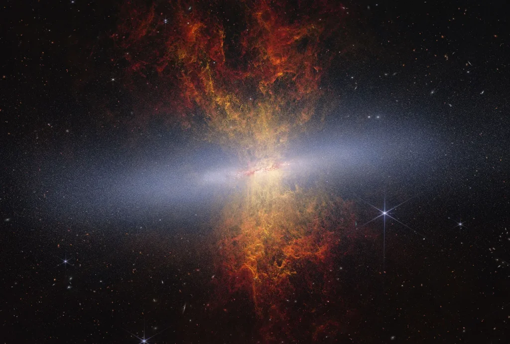 Composite image of edge-on spiral starburst galaxy Messier 82 as observed by NASA's James Webb and Hubble space telescopes. Hourglass-shaped plumes of gas are shooting outward from above and below a bright blue-white, disk-shaped center. The plumes are yellow near the galaxy’s bright center, denoting areas of ionized hydrogen gas as observed by Hubble, and gradually become redder as you move farther away. Messier 82 is set against the black background of space, which has many distant galaxies that appear as small white and orange spirals, ovals, and points of light. Toward the right of Messier 82 is a blue-white star with eight-pointed diffraction spikes that are characteristic of Webb.