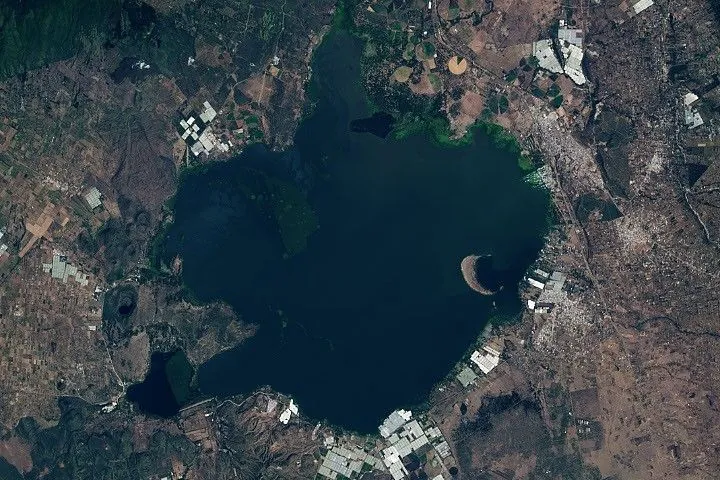 The second image in the pair shows significant encroachment of water on communities, farmland, greenhouses, and infrastructure surrounding the lake by 2026. This image also shows significantly more green mats of water hyacinth in the northern part of the lake.