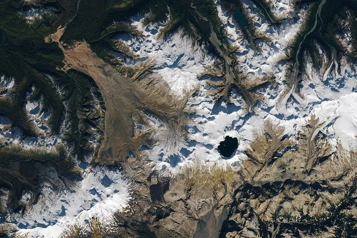 A satellite image shows part of Alaska’s Katmai National Park. A chain of snowy mountain peaks stretches from the bottom left corner to the right side of the image. The Valley of Ten Thousand Smokes is a long, tan-colored feature runs toward the northwest in the upper-left.