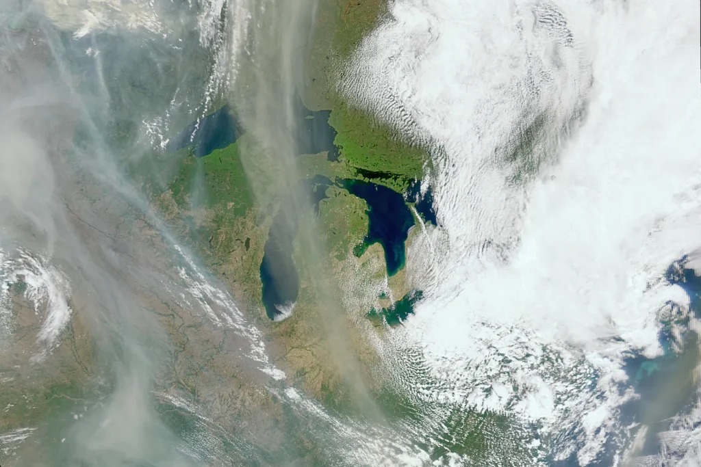 The image is a satellite view. Bright white clouds cover the right half of the image, where little to no land is seen below it. On the left side of the image, gray-tan wispy smoke streaks the image, traveling from top to bottom. Beneath the smoke, green and brown land can be seen, as well as the dark blues of large lakes: The Great Lakes.