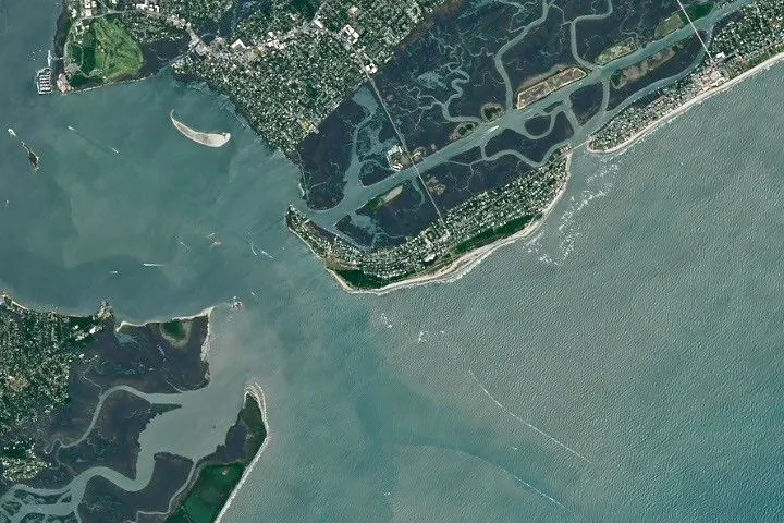 A satellite image shows Sullivan’s Island, South Carolina, with a sandy shoreline along the Atlantic Ocean to the south, development across the interior, and green marsh and tidal inlets along its north side.