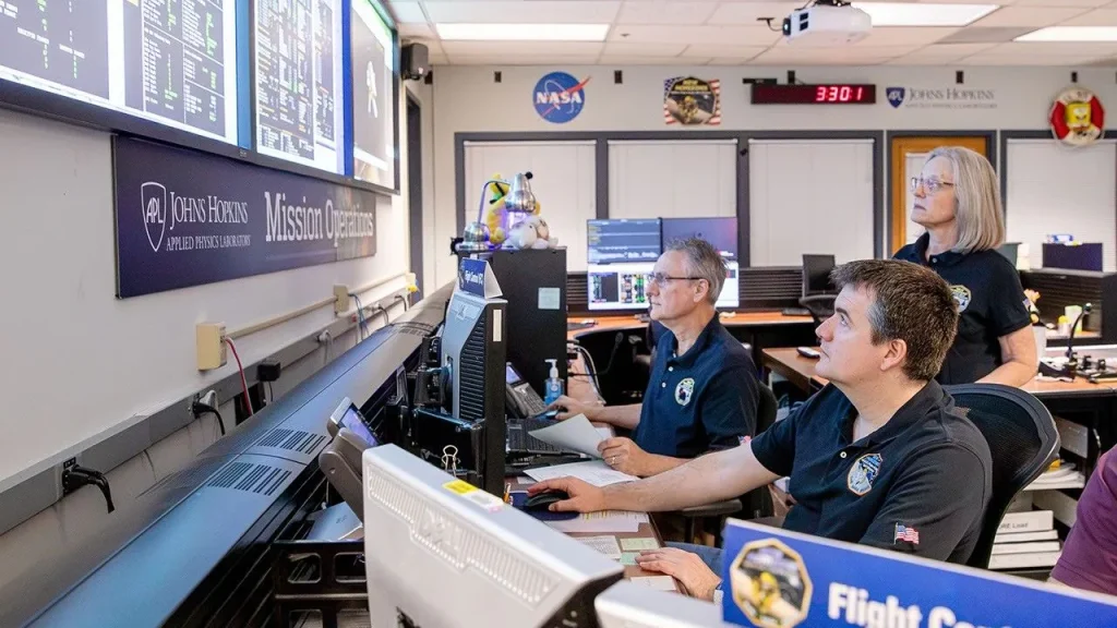 Flight operators at computers in a mission control center monitor spacecraft data on large wall displays.