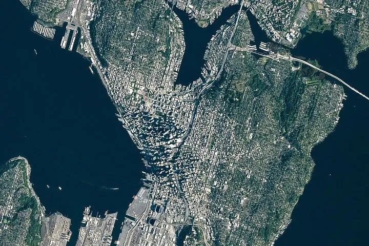 A top-down photo focuses on downtown Seattle, where tall buildings cast long shadows. Green neighborhoods and parks surround the dense urban development, and large waterbodies lie to the east and west.