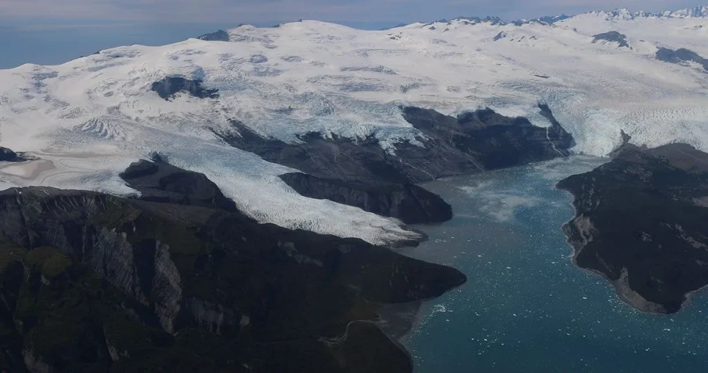 Amid a rugged landscape, a snow-white glacier spills into dark blue water.