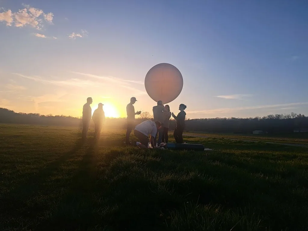 A photograph shows several people in silhouette standing in a large field in front of the setting Sun, gathered around a large, round, white balloon floating just above their heads against a blue sky.