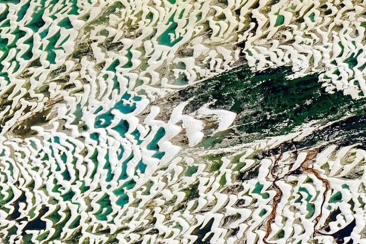 The various blue and greens of the lagoons are more visible in this view of a portion of the dune field. A small river with brown water is visible running northward through the dune field.