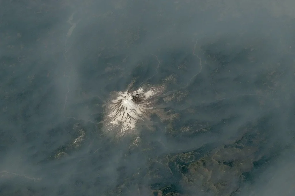 An ice-capped volcano stands at the center of the image, its surrounding terrain blanketed in brown-gray smoke.