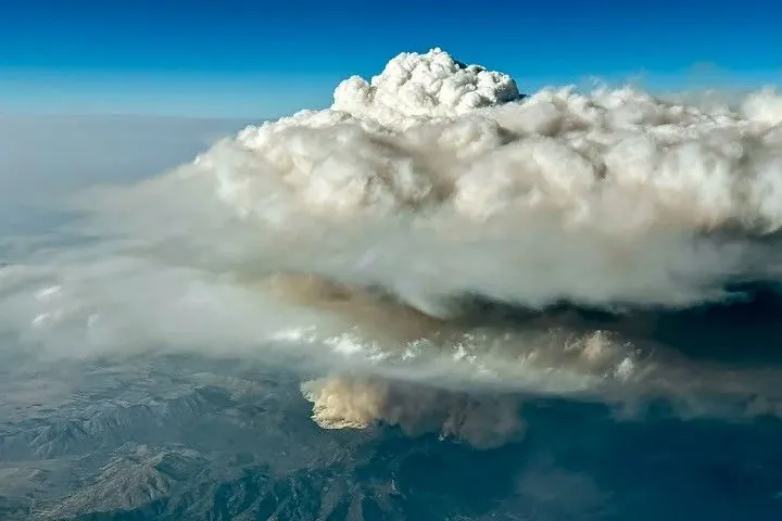 An aerial image shows a thick, puffy white cloud rising high above a patch of darker smoke visible near a surface of variable mountainous terrain.