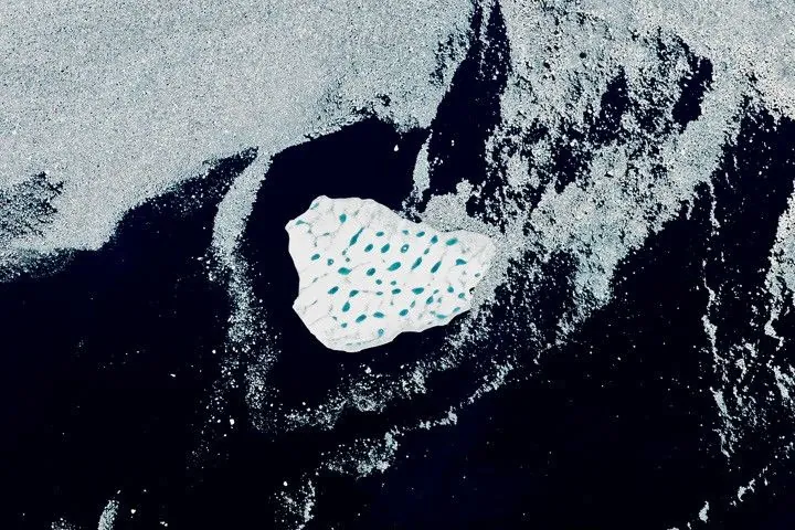 A detailed view shows the iceberg's bright white surface pocked with light blue meltwater ponds. Broken pieces of sea ice float around the iceberg in otherwise dark blue ocean water.