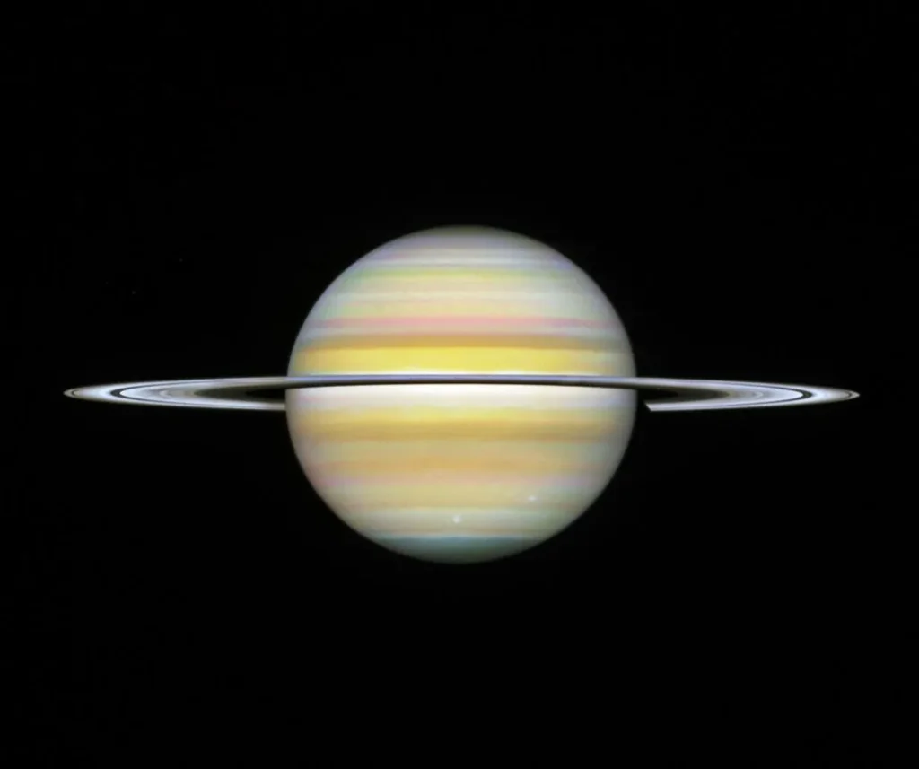 A straight-on view of Saturn, a globe with pale yellow horizontal bands at the equator and orange and pink at the mid-latitudes. Some bands towards the north and south pole have a light blue hue. There are prominent horizontal rings circling at the equator.