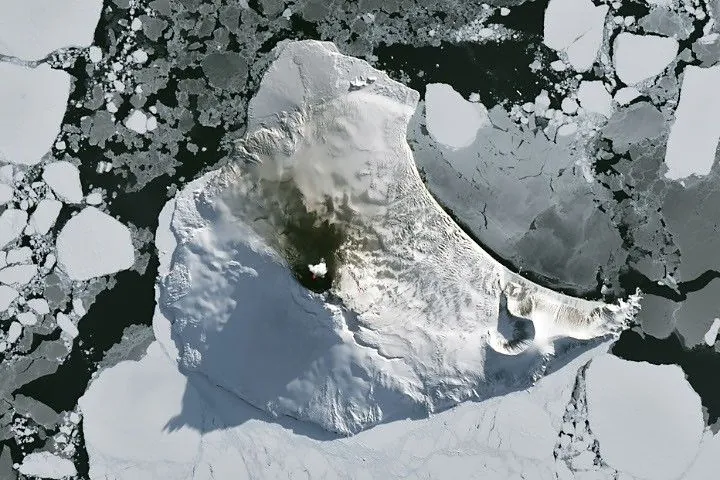 A small ice- and snow-covered island with an active volcanic crater at its center is surrounded by drifting pieces of sea ice. A thermal signal and small plume appear in the crater, and ash darkens the snow on the volcano’s northern slopes.