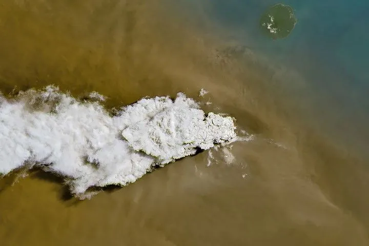 A white volcanic plume streams left over a tan ash cloud that fills most of the image. A small green island and blue water are visible in the upper right.