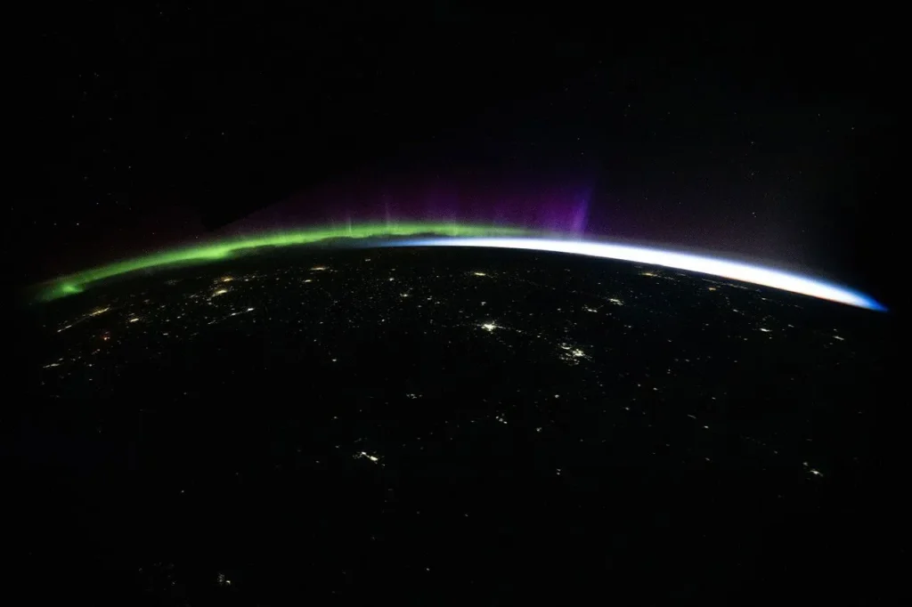 Green and purple aurora and white airglow arc above Earth’s limb and the scattered city lights of Central Asia in a nighttime photo taken from the International Space Station.