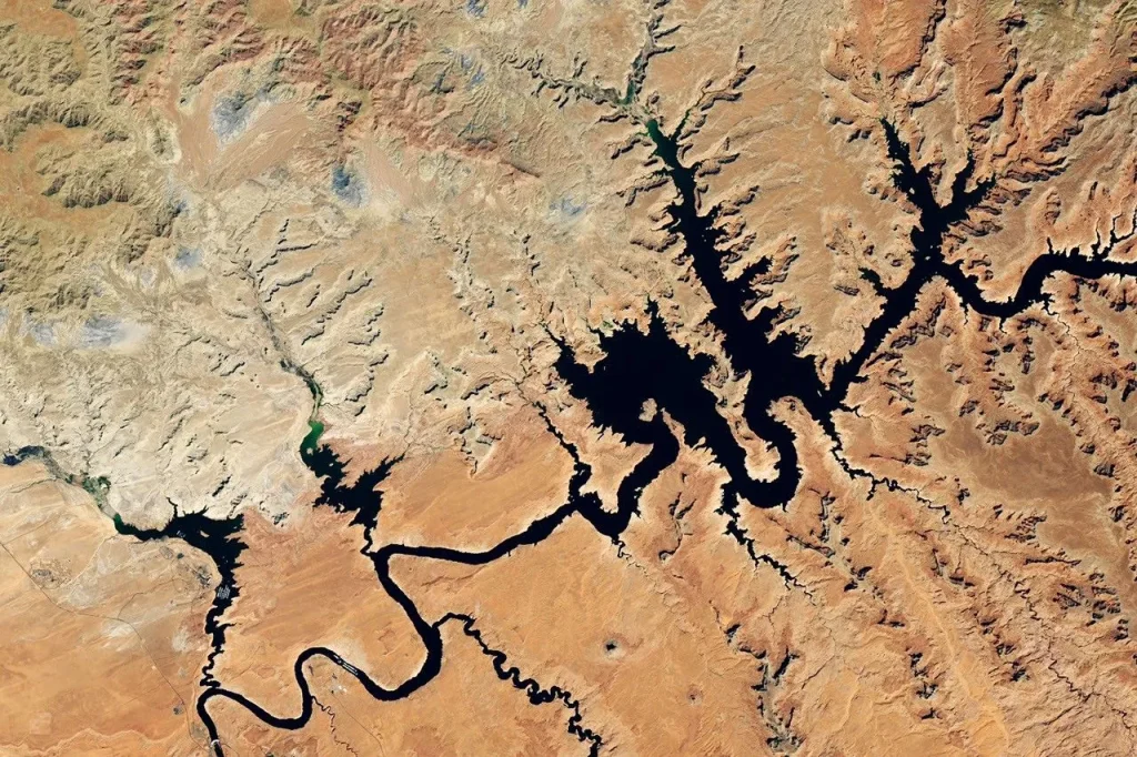 Water in Lake Powell is at record-low levels and looks like a wide river winding through a light orange desert landscape. The Glen Canyon Dam and the town of Page, Arizona, are visible in the lower left.